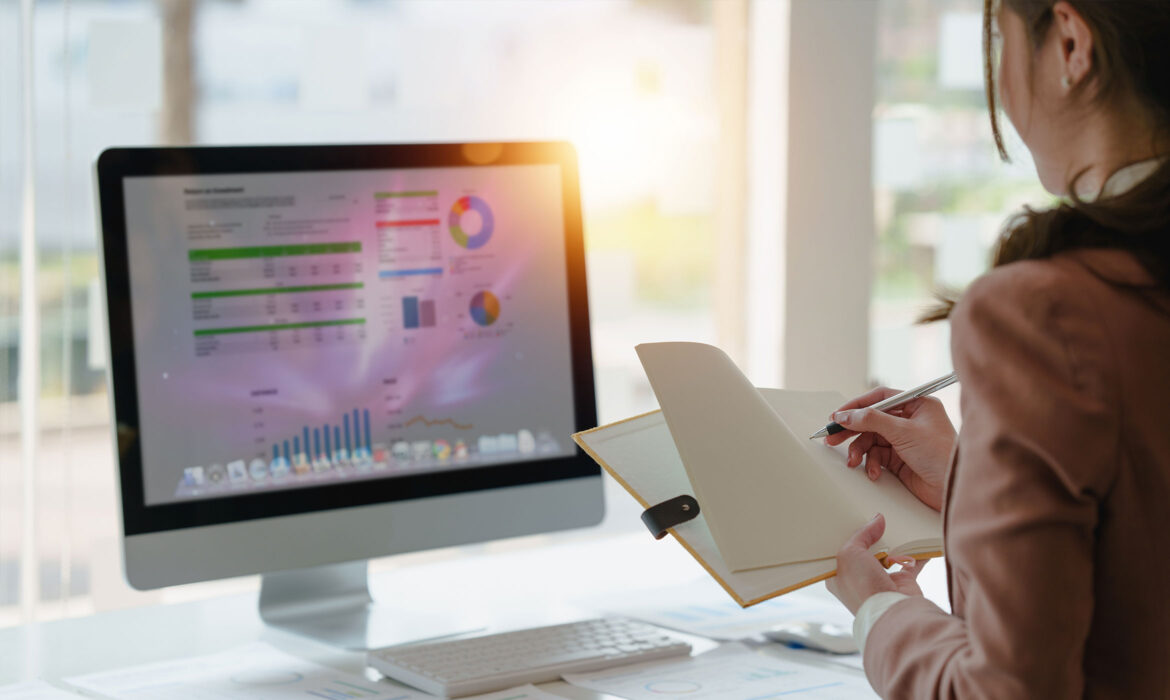 Femme prenant des notes dan sun carnet devant un ordinateur présentant des graphes et des statistiques